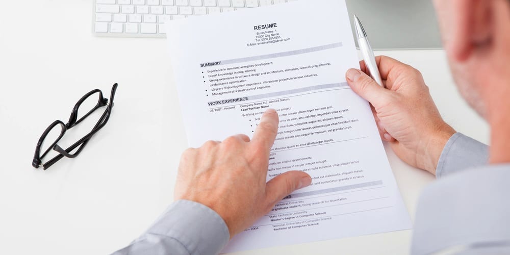 A man holding a printed resume, a glasses on the left of the table and on his right hand he holds a pen