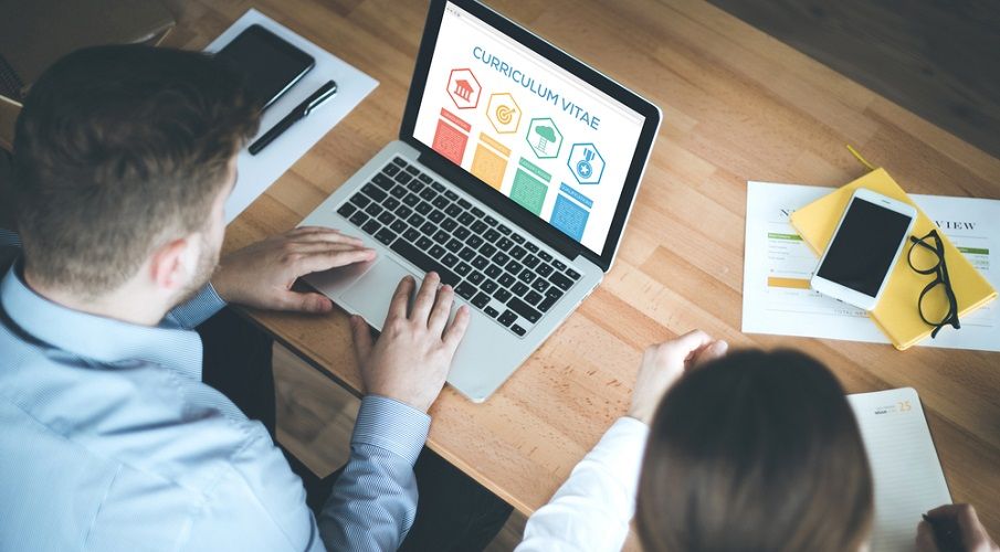 A men on a laptop with a curriculum vitae screen and a woman on his right writing on a paper