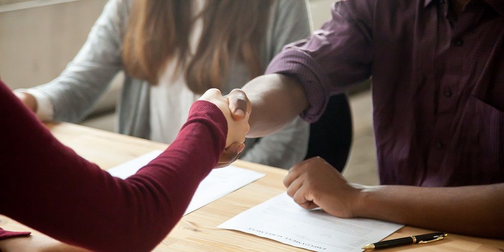 A table with two people shaking hands and other person on the left. Some papers on the table.