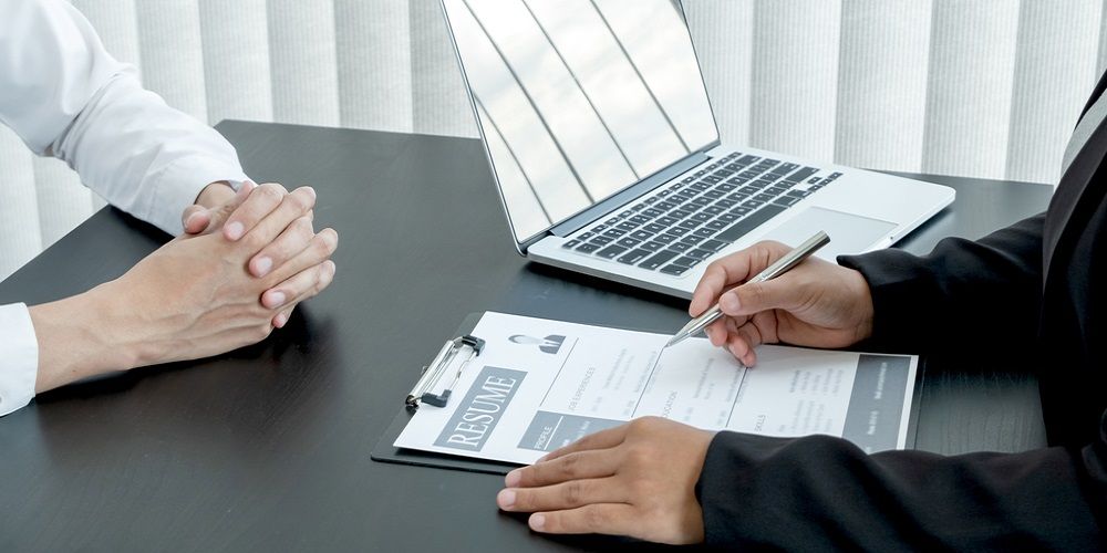 Two people with hands on the table and one of them has a resume and a laptop