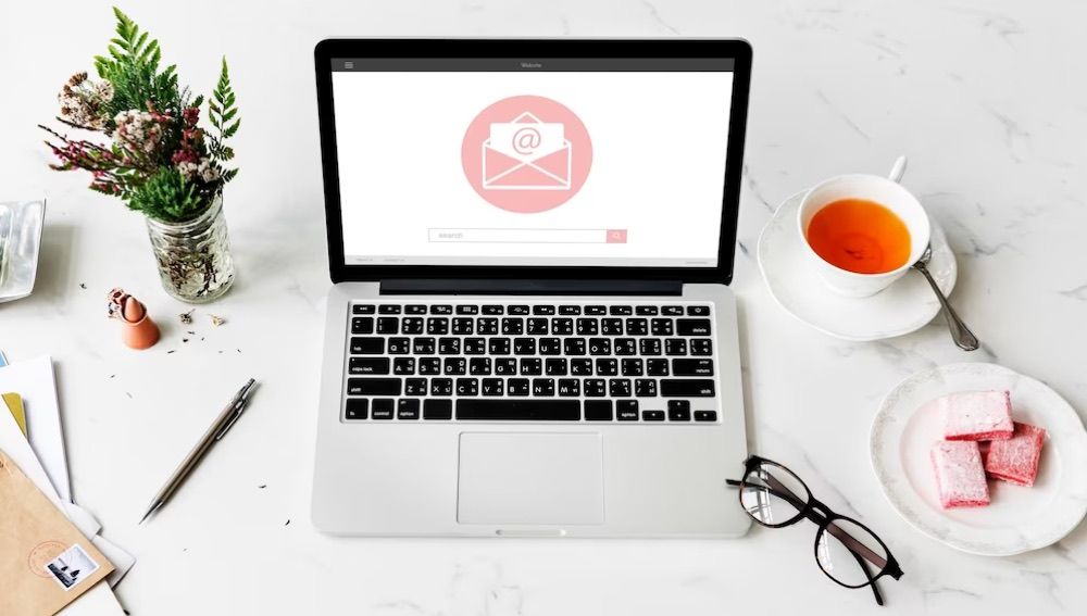 Laptop on a marble desk showing an email icon on screen, surrounded by tea, a plant, and reading glasses
